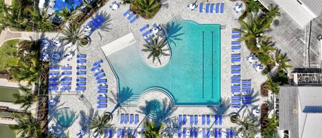 An aerial view of a resort with a central swimming pool surrounded by sun loungers and palm trees.