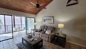 Un salon spacieux doté d’un ventilateur de plafond, d’une grande fenêtre, d’un canapé en cuir marron, d’une table basse et d’une table d’appoint avec une lampe.