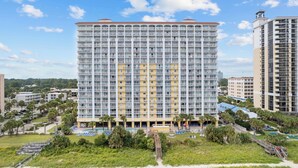 A high-rise hotel with multiple balconies, a swimming pool, and a landscaped area with palm trees. - 15th Floor Oceanfront Condo with Beach, 4 Pools, 2 Hot Tubs & a Lazy River (Myrtle Beach)