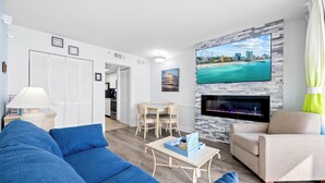 A modern living room with a blue sofa, a beige armchair, a coffee table, a TV mounted on a stone wall, and a fireplace. - 15th Floor Oceanfront Condo with Beach, 4 Pools, 2 Hot Tubs & a Lazy River (Myrtle Beach)