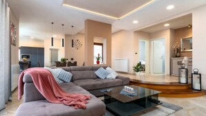 Una sala moderna con un sofá seccional gris, una mesa de centro de vidrio y una manta rosa. En el fondo, se distingue un comedor con una mesa y sillas blancas.