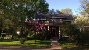 Une maison à deux étages avec un porche à l’avant, entourée d’une végétation luxuriante et d’arbres en fleurs.