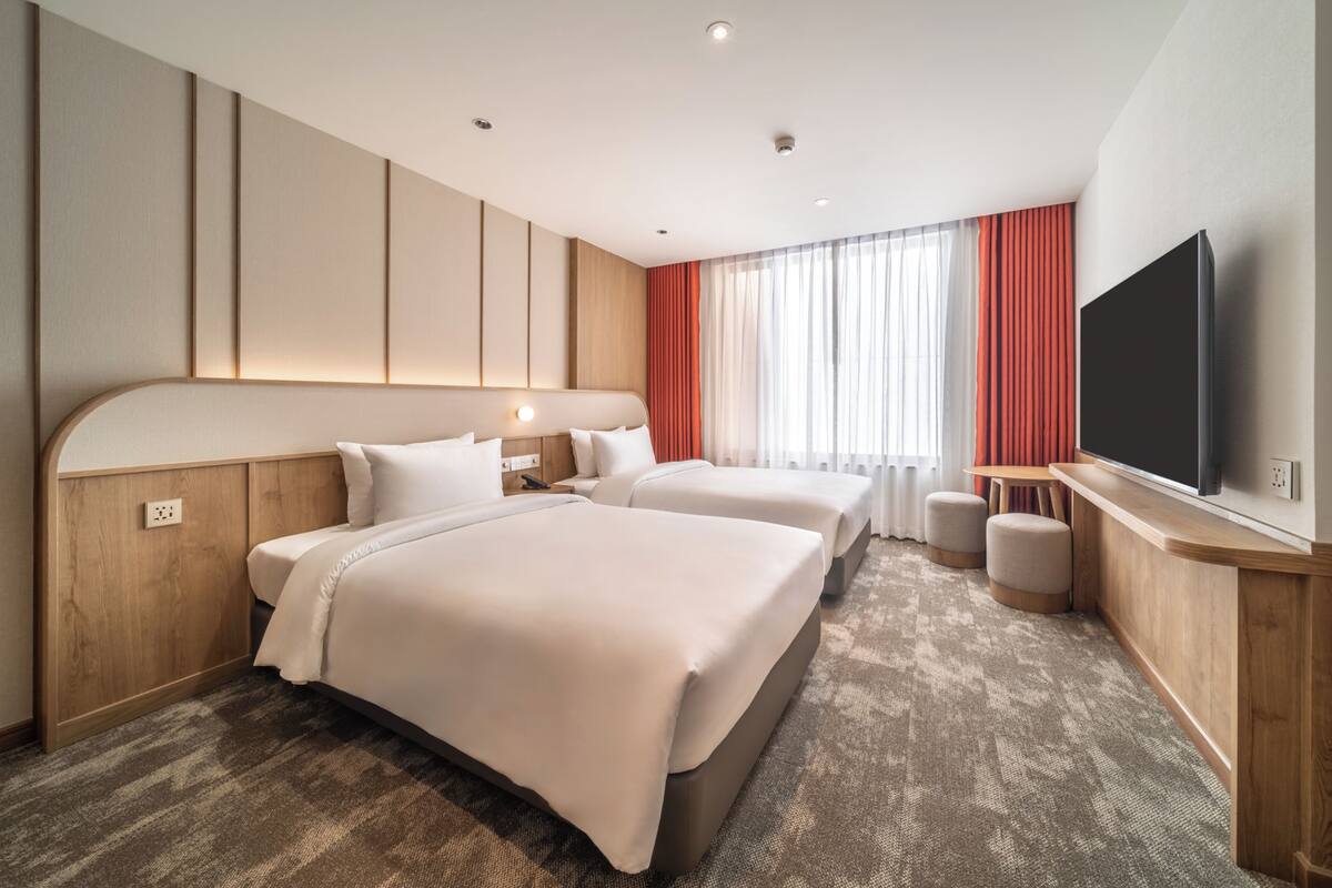 Ein Hotelzimmer mit zwei Betten, einem Fernseher, einem Schreibtisch und einem großen Fenster mit Vorhängen.