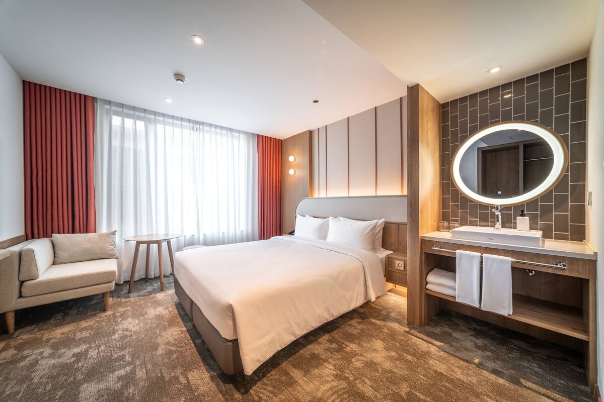 Ein Hotelzimmer mit einem großen Bett, einem Nachttisch, einer Couch, einem runden Spiegel und einem fest eingebauten Waschbecken.