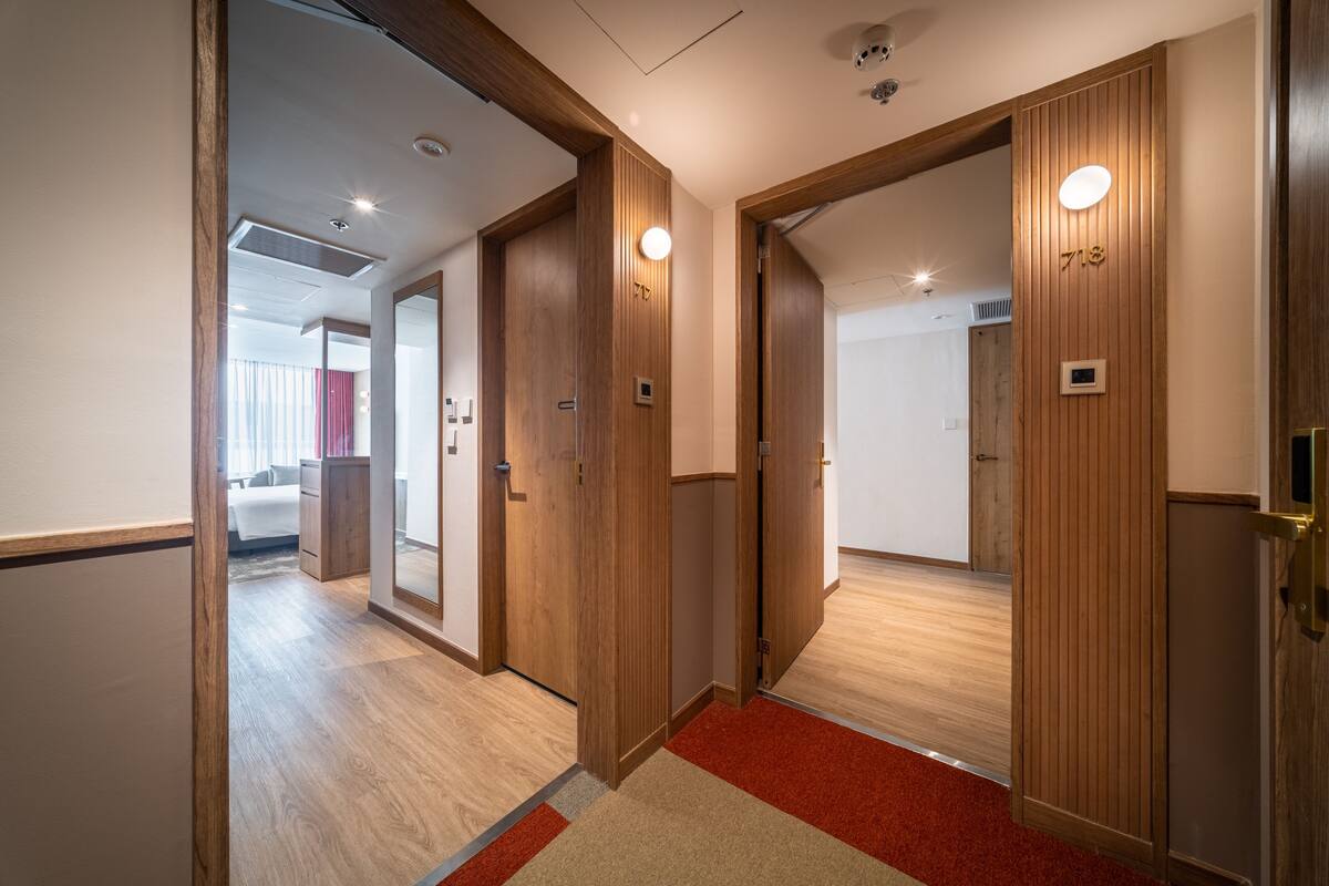 Ein Flur mit Holztüren, einem roten Teppich und einem sichtbaren Zimmer im Hintergrund, das ein Bett zeigt.