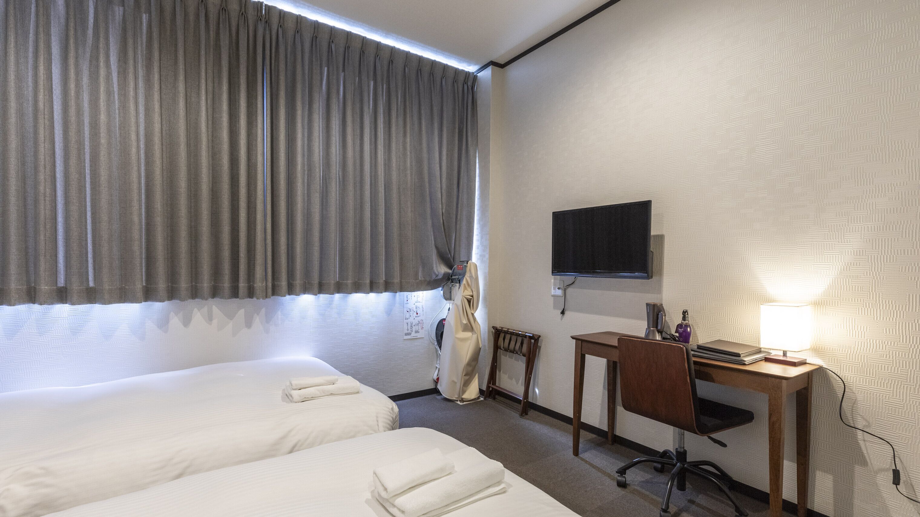 A hotel room with two beds, a desk with a lamp, a television, and a window with curtains.