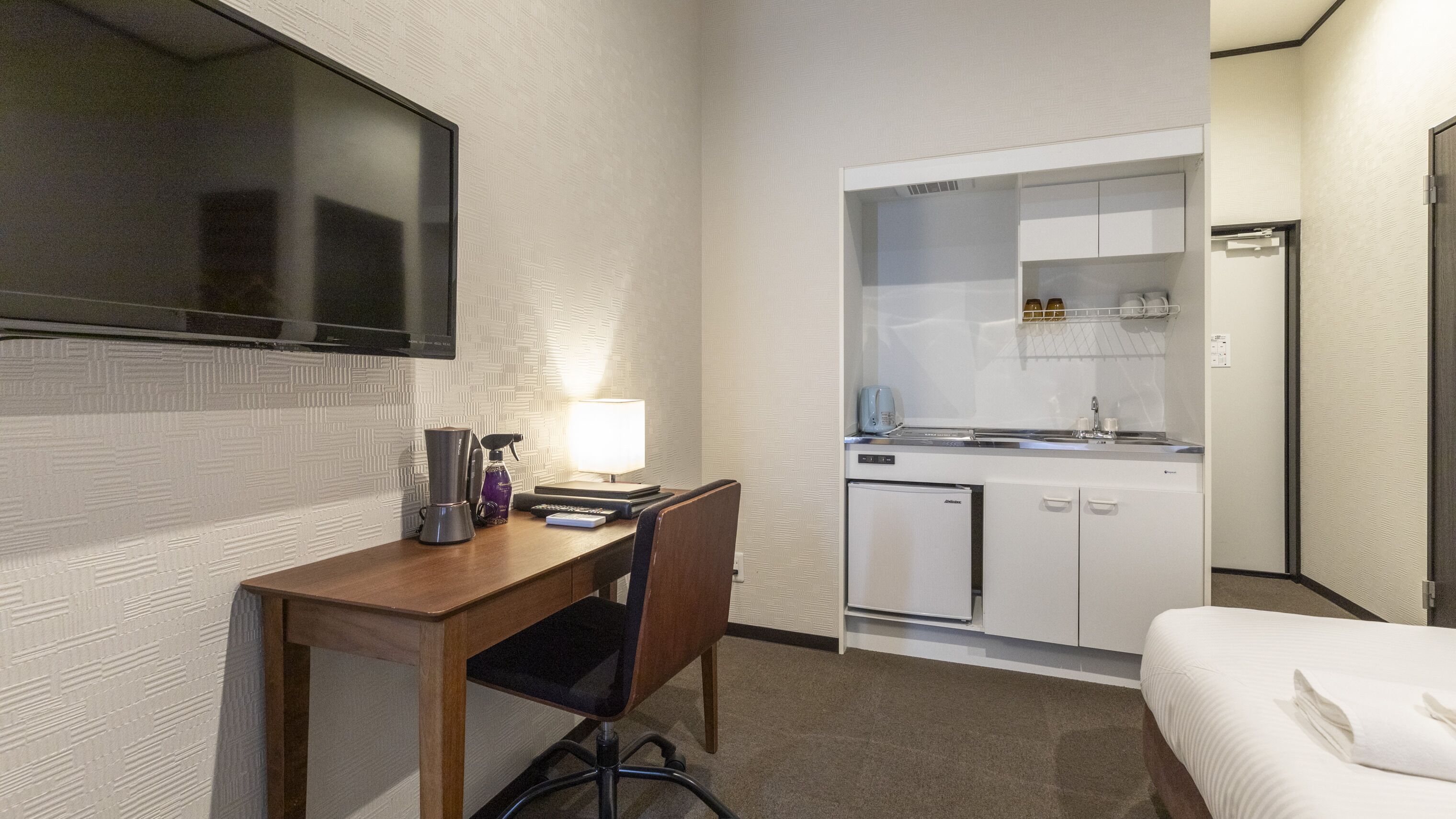 Habitació d'hotel petita amb un llit, una taula de treball amb una cadira, una cuineta i un televisor fixat a la paret.