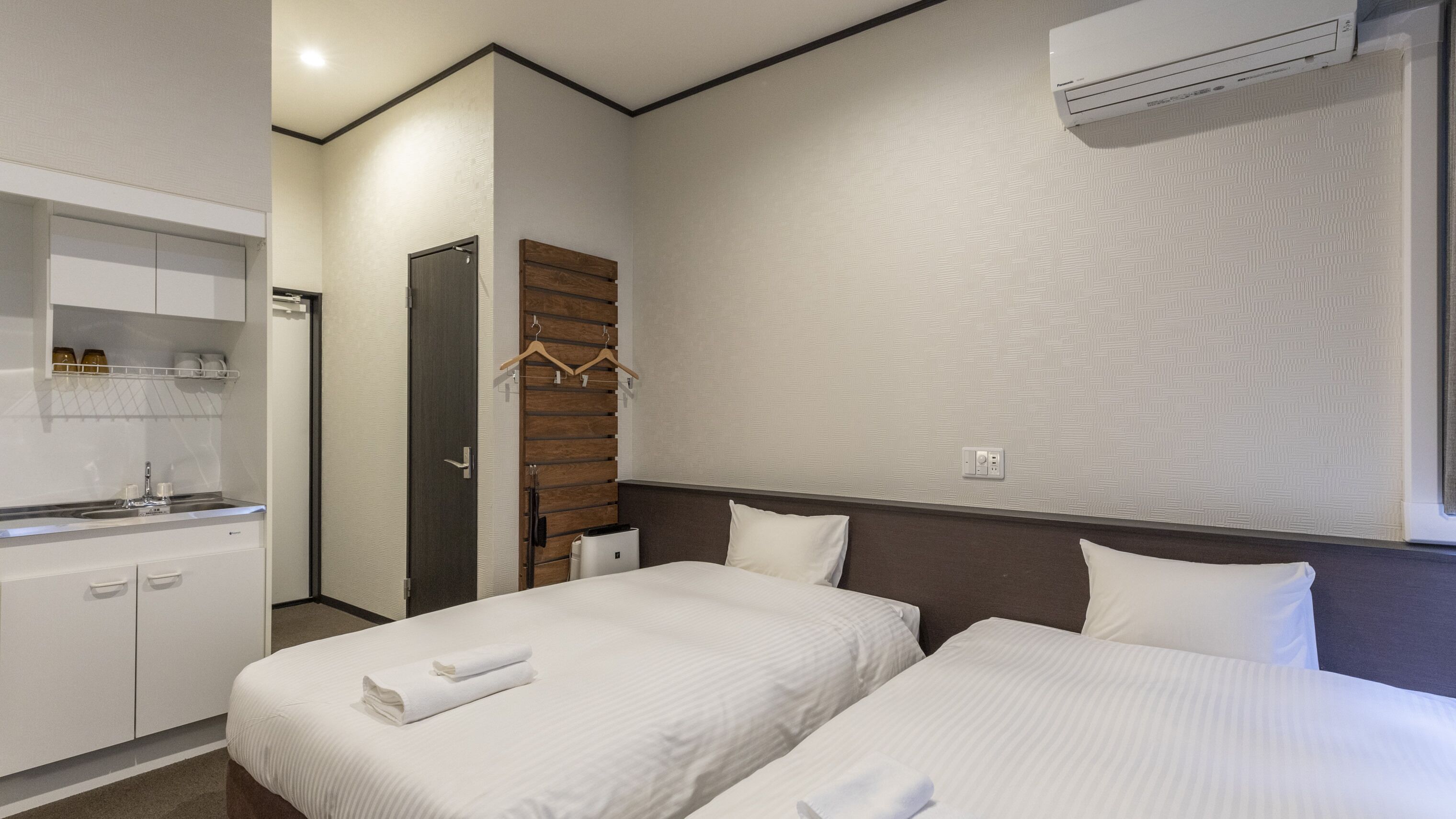 A hotel room with two beds, a small kitchenette, a wooden panel wall, and an air conditioning unit.
