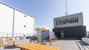 An outdoor seating area with wooden tables and chairs, air conditioning units, and a large building with a sign reading "OSAKA PORT HOTEL L'ESPOIR". - OSAKA PORT HOTEL L'ESPOIR (Osaka)