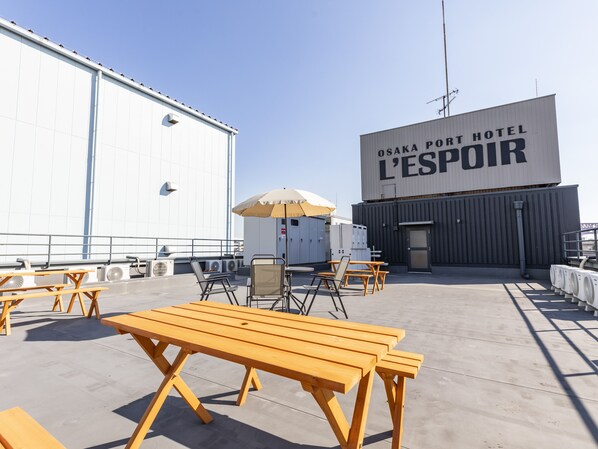An outdoor seating area with wooden tables and chairs, air conditioning units, and a large building with a sign reading "OSAKA PORT HOTEL L'ESPOIR". - OSAKA PORT HOTEL L'ESPOIR (Osaka)