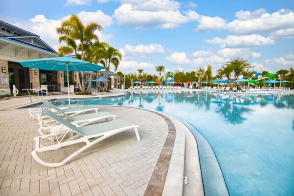 A swimming pool with lounge chairs and umbrellas, surrounded by palm trees and a building with a blue roof.