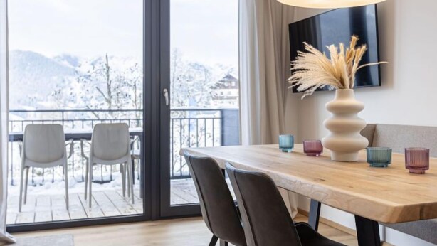 A modern dining area with a wooden table, chairs, and a large window offering a view of snow-covered mountains.