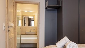 A modern hotel room with a wooden door, a bathroom with a sink and mirror, a bed with white linens, and a dark-colored wardrobe.