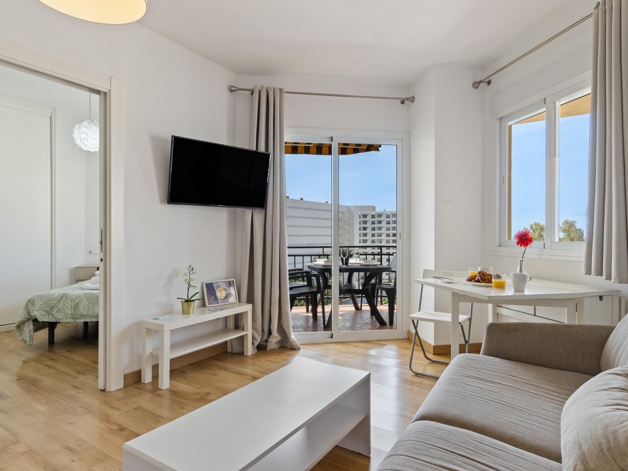 A modern living room with a flat-screen TV, a sofa, a coffee table, a balcony with outdoor furniture, and a dining area.