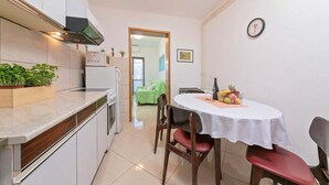 Eine kompakte Küche mit Mikrowelle, Herd und eingebautem Backofen. Ein Esstisch mit Stühlen, eine Uhr an der Wand und eine kleine Pflanze auf der Arbeitsplatte sind vorhanden.