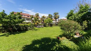 Ein gepflegter Garten mit vielfältigen Pflanzen und Bäumen, ein klarer, blauer Himmel und im Hintergrund ein Gebäude mit roten Ziegeldächern.