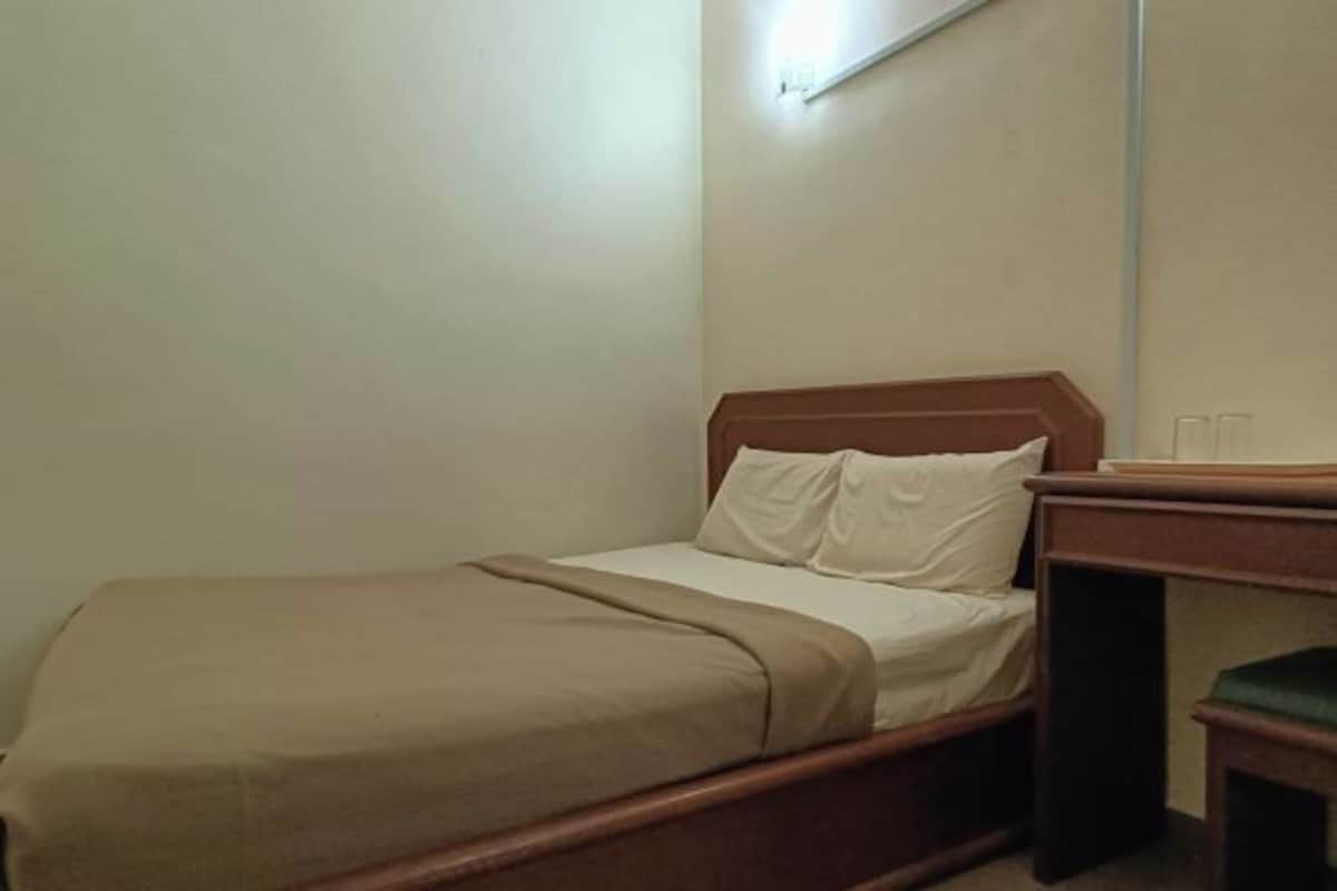 Una habitación pequeña con cama individual, cabecera de madera, mesita de noche y una silla verde.