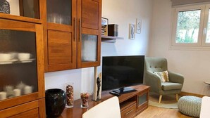 Un salon avec des meubles de rangement en bois, un téléviseur à écran plat, un fauteuil gris et un pouf rond.