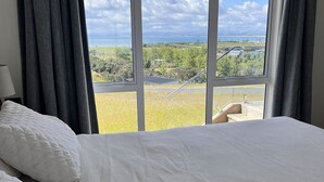 A bedroom with a large bed, a view of the outdoors, and a window with curtains. - Karikari Vista (Karikari Peninsula)