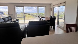 A modern living room with a large sliding glass door leading to a balcony. There is a black leather sofa and a dining table with chairs. - Karikari Vista (Karikari Peninsula)