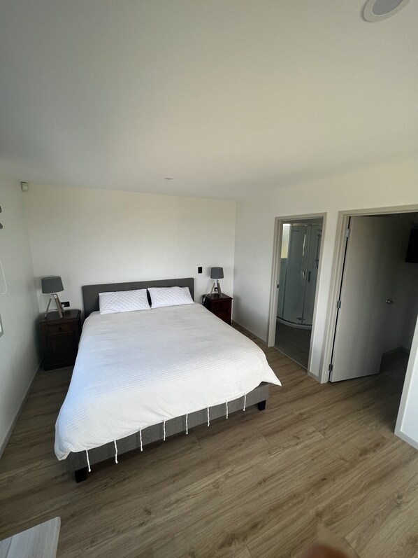 Un dormitorio con una cama grande, piso de madera, una mesita de noche con una lámpara y una puerta que da a otra habitación.