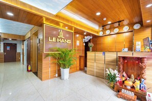 Un espace d’accueil d’hôtel avec un comptoir de réception en bois, une grande enseigne indiquant « Le Hang Hospitality & Hotel Management », un extincteur et une plante en pot.