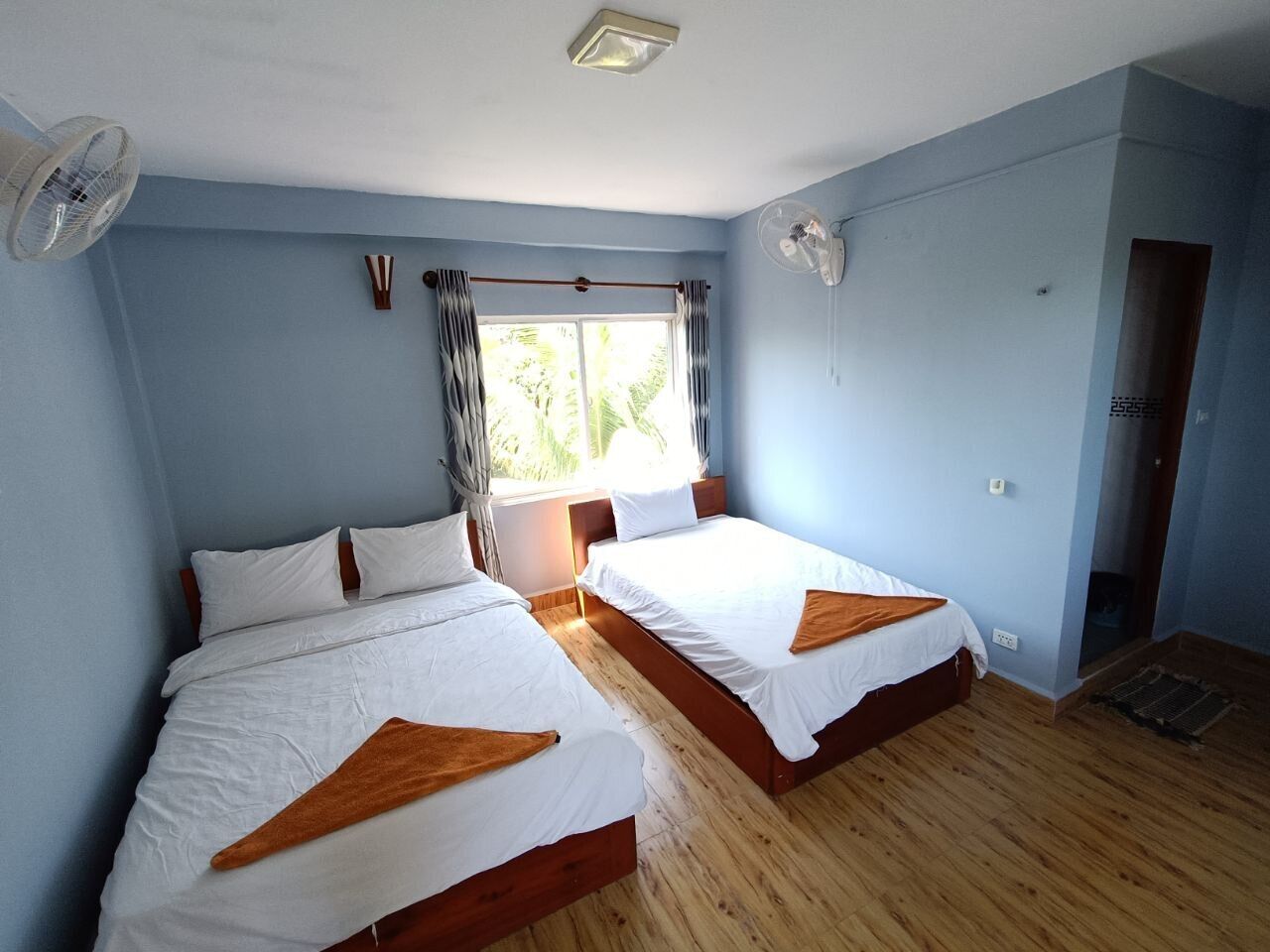 A room with two beds, wooden flooring, a window with curtains, and a ceiling fan.