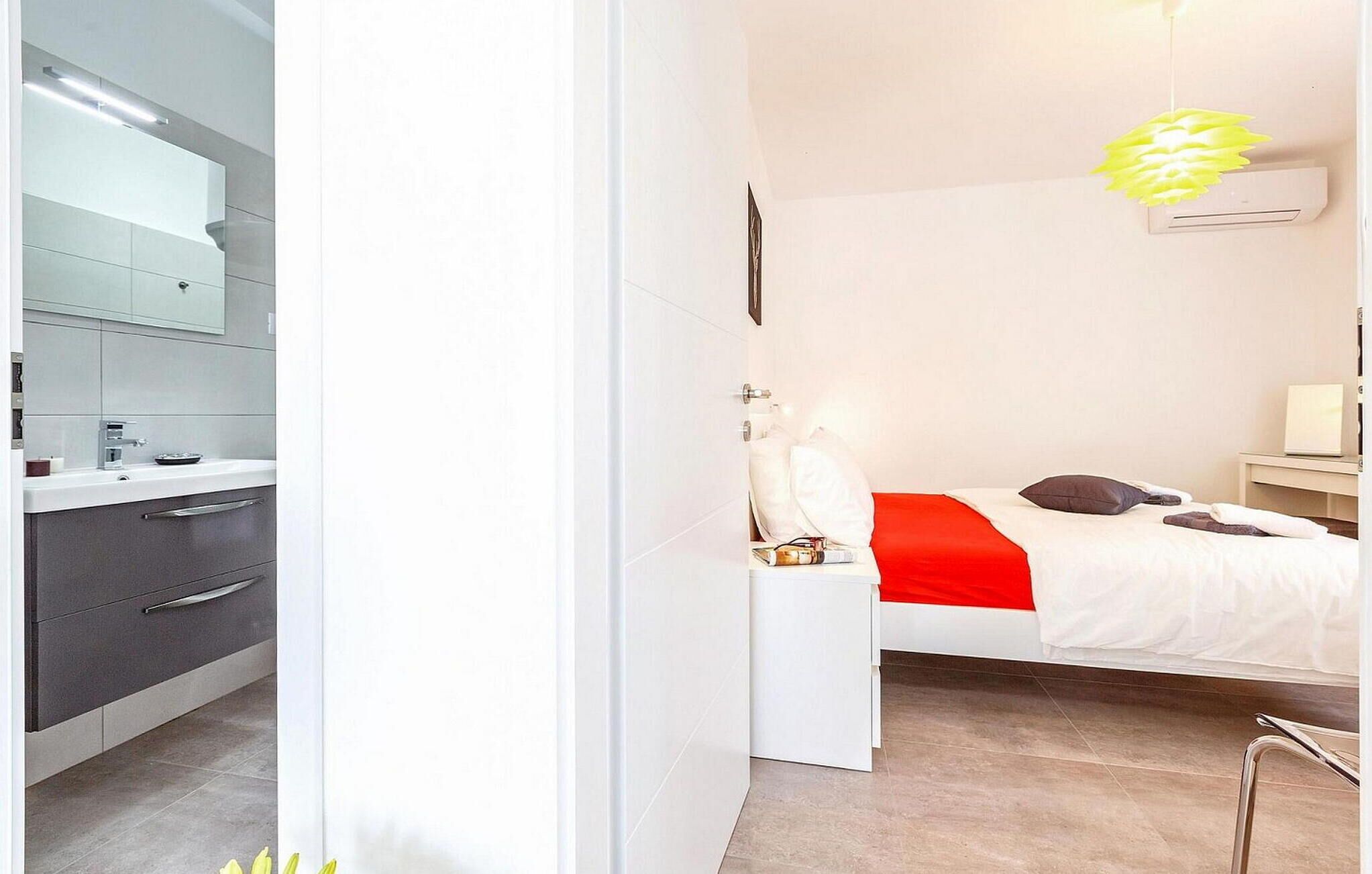 Habitación de hotel moderna con cama blanca, mesita de noche, espejo y baño con lavamanos y armarios.