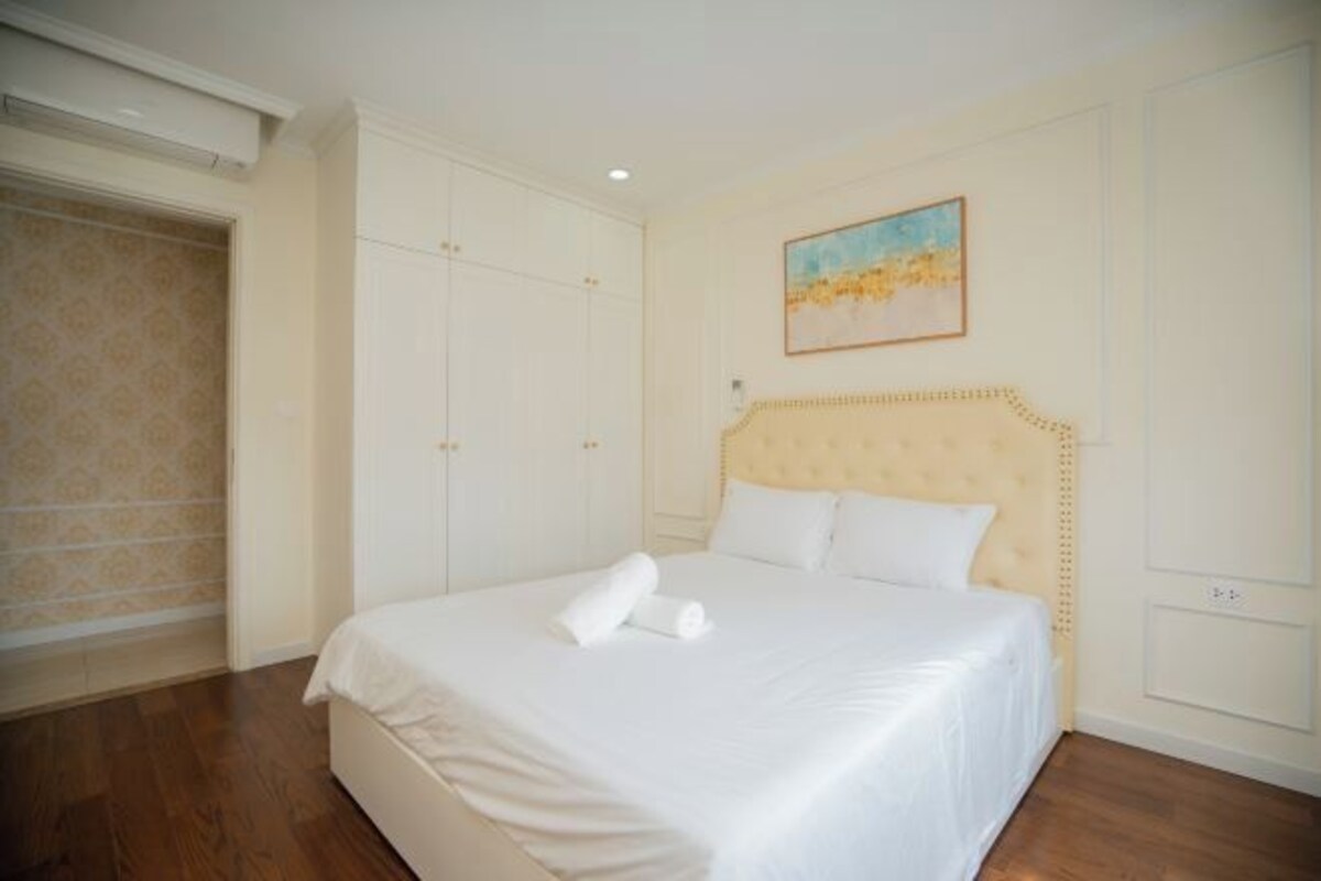 A neatly made bed with a white duvet and a rolled towel, a headboard with a decorative pattern, and a painting on the wall.