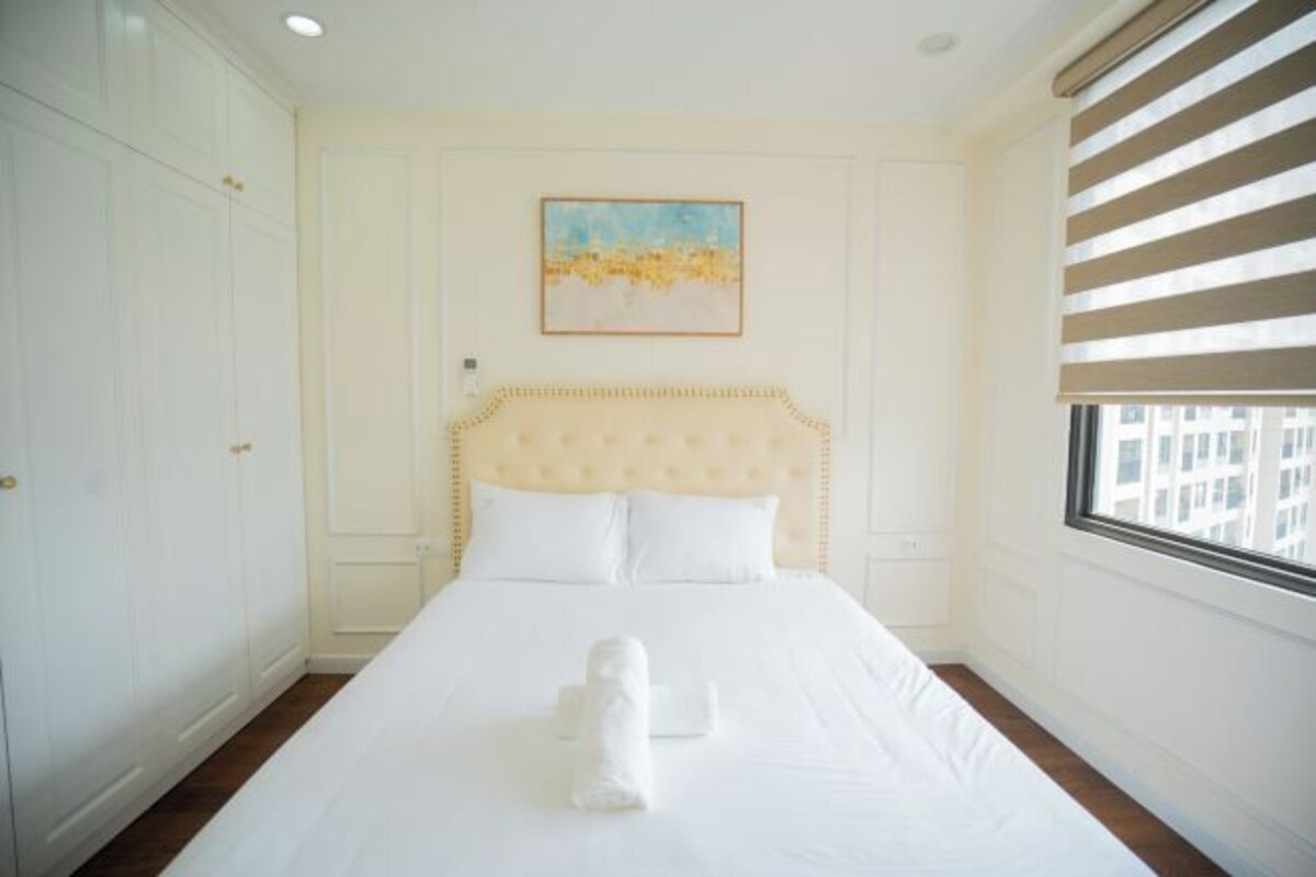 A hotel room with a large bed, a headboard, a painting on the wall, and a window with blinds.