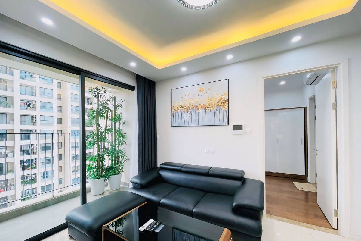 Un salon moderne avec un canapé en cuir noir, une table basse en verre et une grande fenêtre donnant sur un quartier résidentiel.