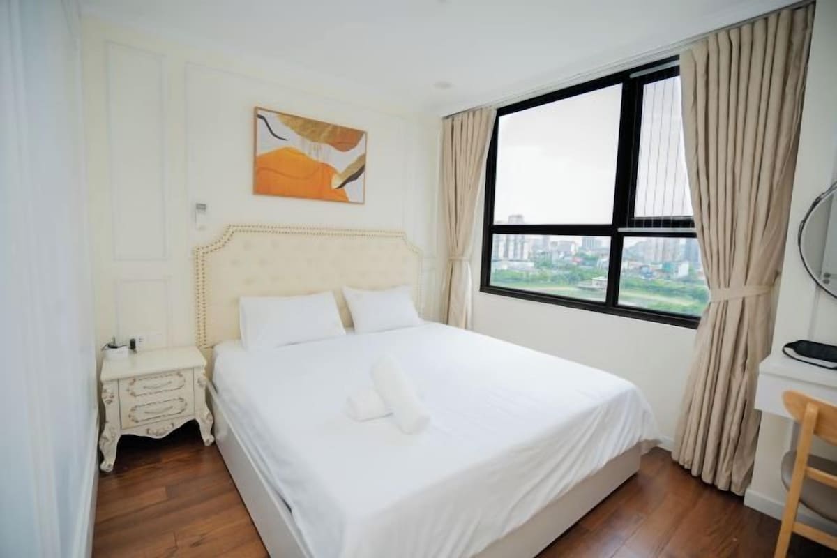 Quarto de hotel com uma cama grande, mesa de cabeceira, escrivaninha e janela com cortinas.