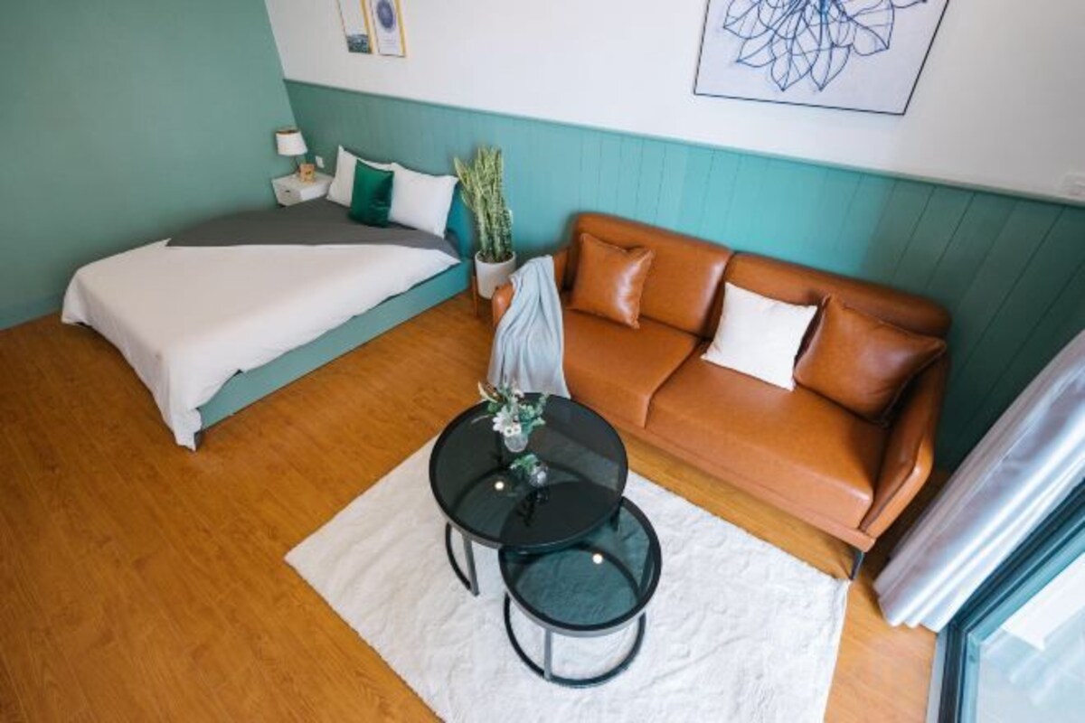 A room with a bed, a brown leather sofa, a small table with a vase, and a window with curtains.
