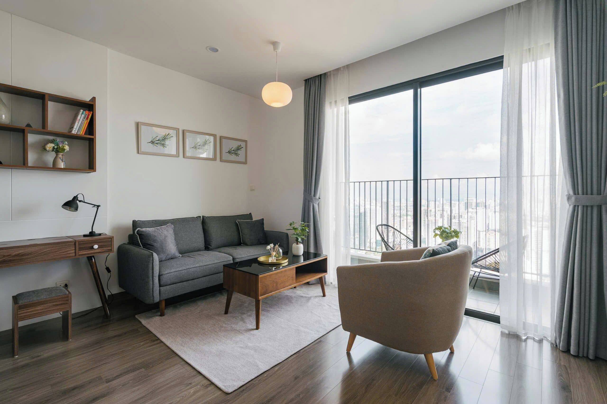 A modern living room with a grey sofa, a wooden coffee table, a grey armchair, a bookshelf, and a large window with a city view.