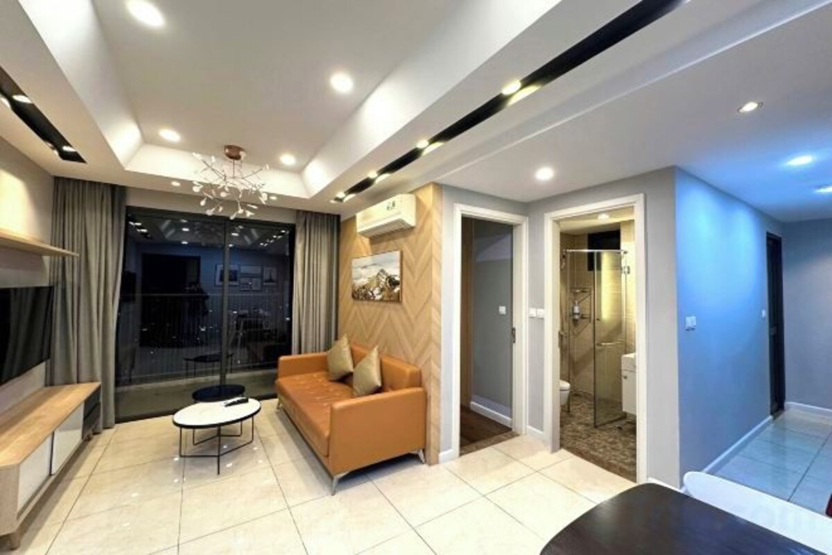 A modern living room with a sofa, a coffee table, and a TV. There is a glass door leading to a bathroom.