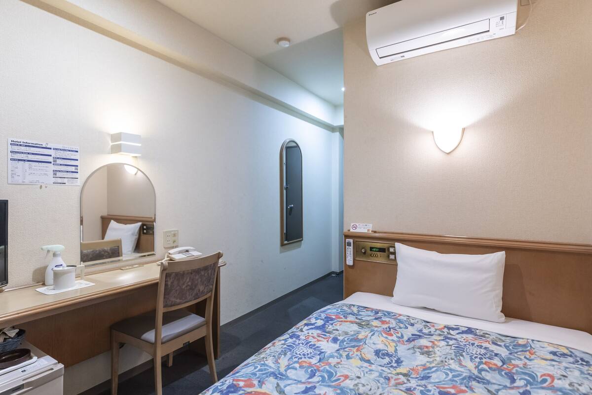 A hotel room with a bed, a desk with a chair, a mirror, an air conditioner, and a wall-mounted control panel.