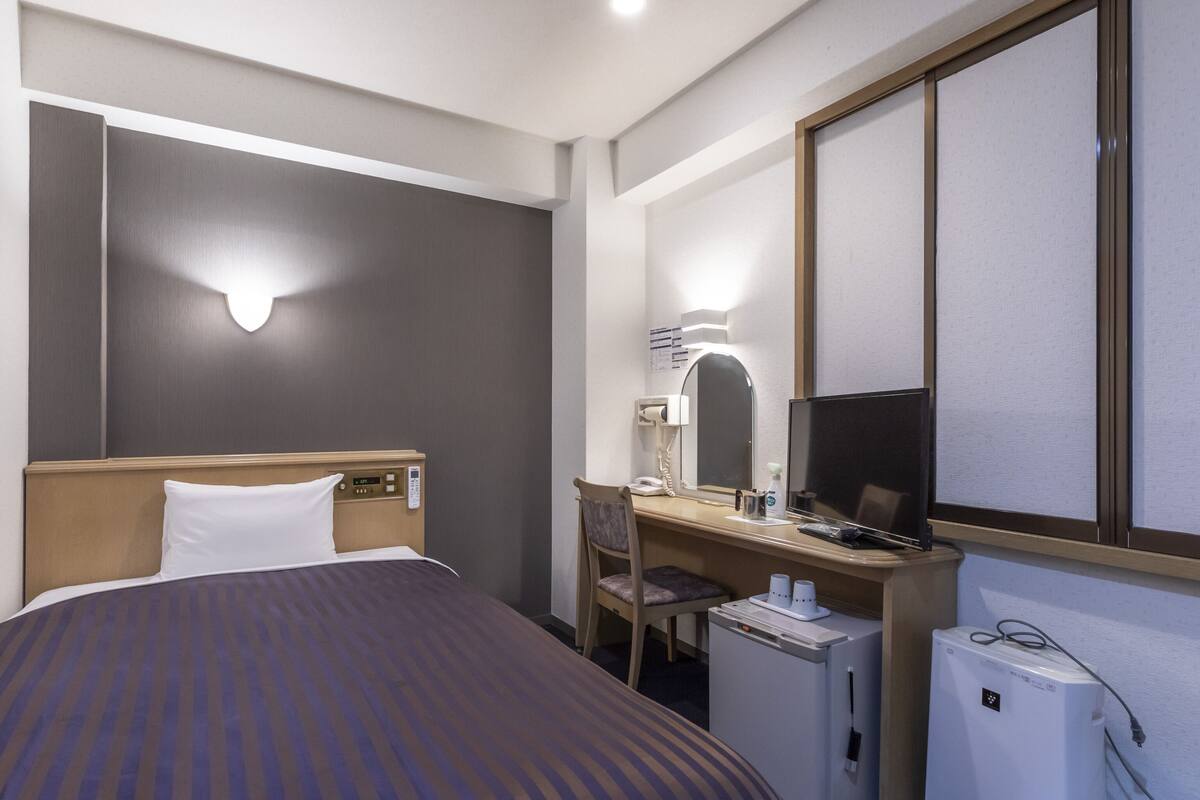 A hotel room with a bed, desk, chair, and a small refrigerator.