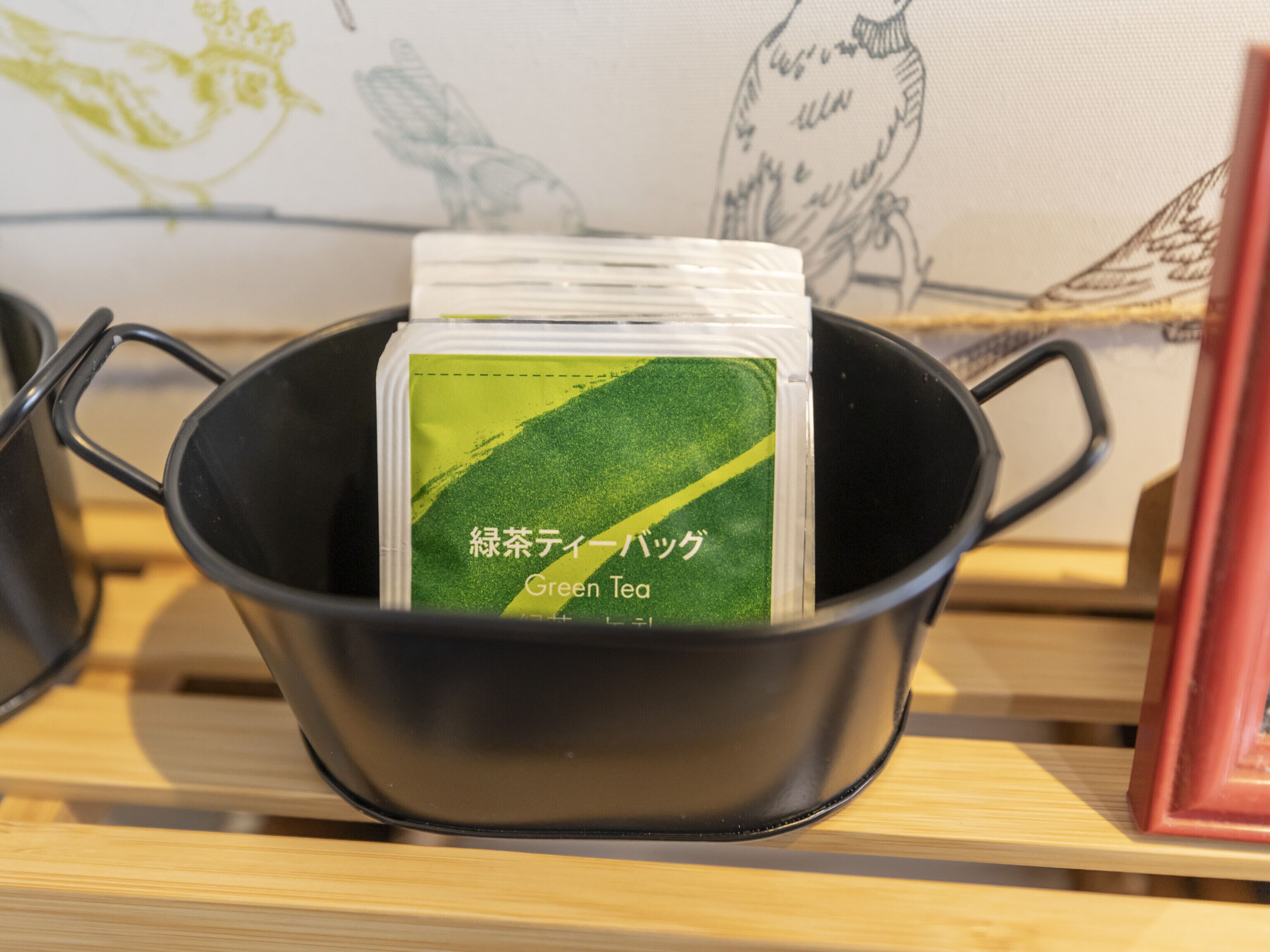 Eine schwarze Keramikdose mit einem Grünteebeutel, der mit 'Green Tea' beschriftet ist, auf einem Holzregal.