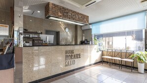A hotel reception desk with a 'HOTEL HORAIRE SAITO' sign, a computer, a cash register, and a window with a view of the outside. - Hotel HORAIRE Saijo (Saijo)
