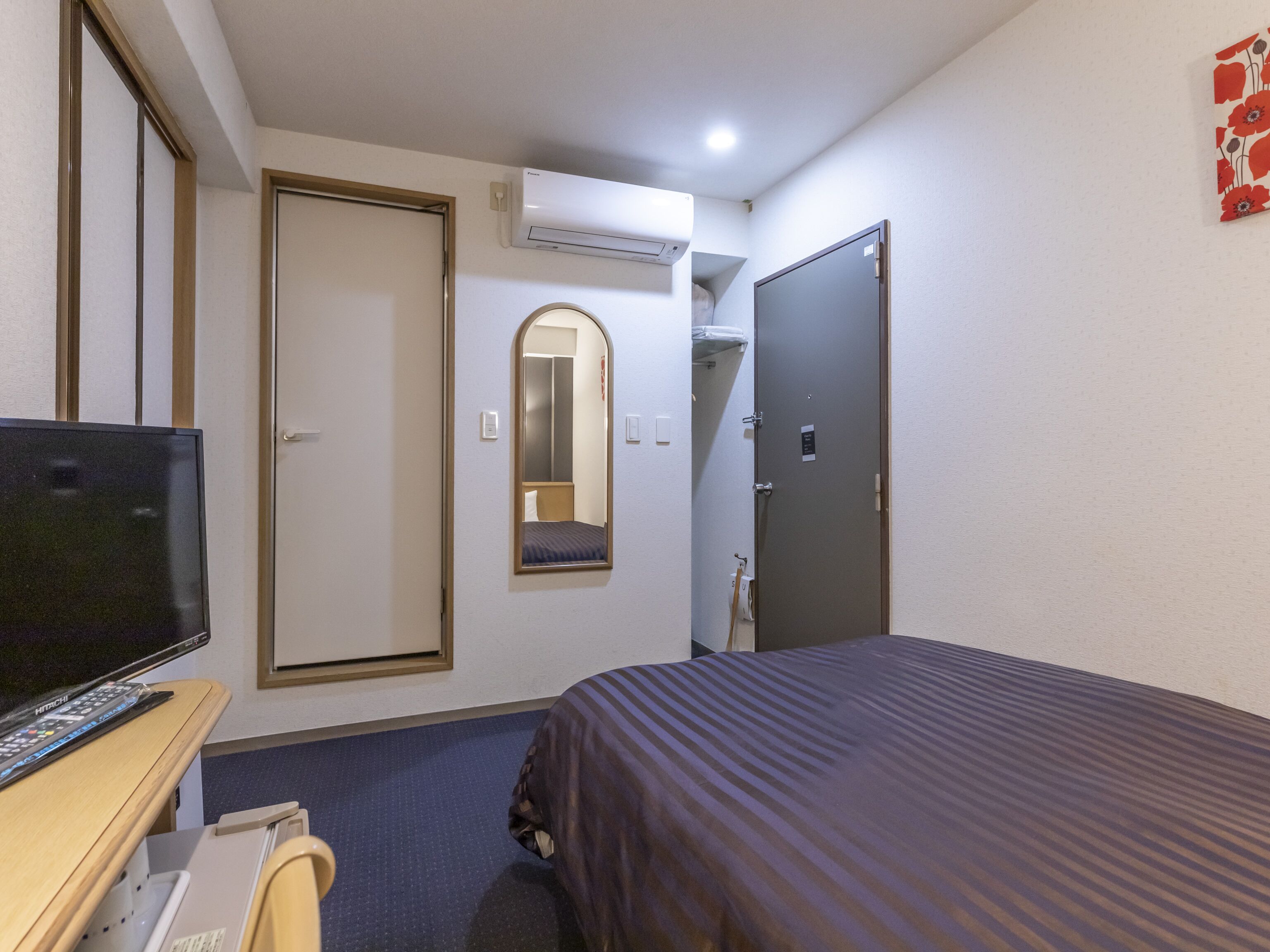 A hotel room with a bed, a desk with a television, a wardrobe, a mirror, and an air conditioning unit.