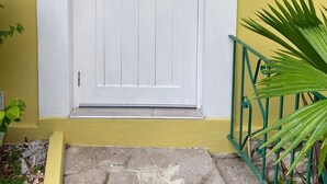 A white door with a brass doorknob and a brass lock, set in a yellow wall with a green metal railing and a potted plant beside it. - 3 BDR Apt at Ramparts Near Sangster Airport (Montego Bay)