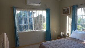 A bedroom with a bed, a standing fan, a small table with a lamp, and a window with curtains. - 3 BDR Apt at Ramparts Near Sangster Airport (Montego Bay)
