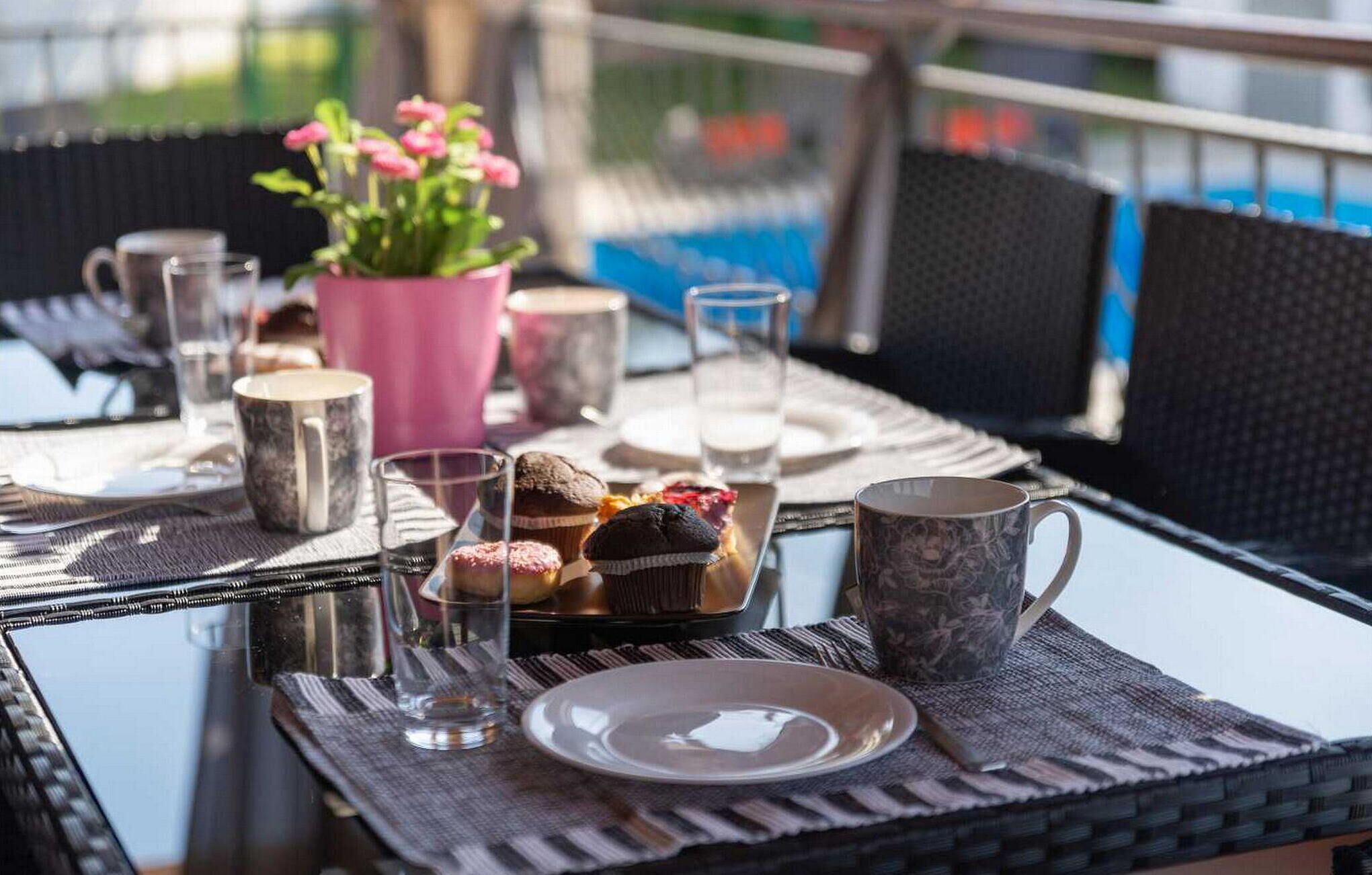 A table set with plates, glasses, a pink flower pot, and a basket of muffins.