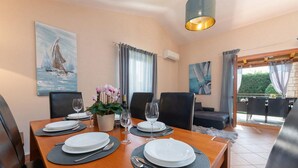 Un comedor con una mesa de madera dispuesta para cuatro personas, rodeada de sillas negras. Hay dos cuadros en las paredes, una planta en maceta con flores y un equipo de aire acondicionado.