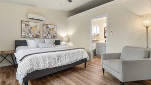 Une chambre moderne avec un grand lit, un bureau et une chaise. On y trouve un ventilateur de plafond, un appareil de climatisation et des œuvres d’art accrochées au mur.