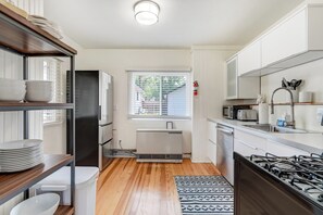 A compact kitchen with a gas stove, white cabinets, a microwave, and a window with blinds. - Sophies Cottage (Colorado Springs)