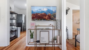 A modern kitchen with wooden flooring, a stainless steel refrigerator, and a wall-mounted TV displaying a mountain landscape. - Sophies Cottage (Colorado Springs)