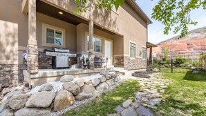 Une maison moderne avec une façade en pierre, un patio doté d’un barbecue et un chemin en pierre menant à l’entrée.