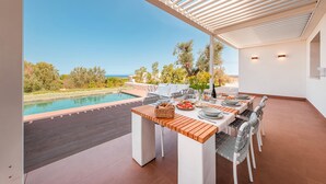 Un área de comedor junto a la piscina con vistas a la piscina y al océano, mesas de madera, sillas blancas y una pérgola.