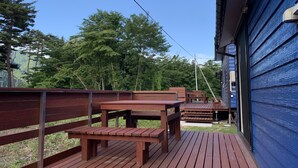 A wooden deck with benches and tables, surrounded by a blue building and a forested area. - Kita-alps BASE Onsenkyo Camp (Omachi)